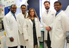Five smiling clinical fellows in white coats standing in the lab. Courtesy NIH flickr